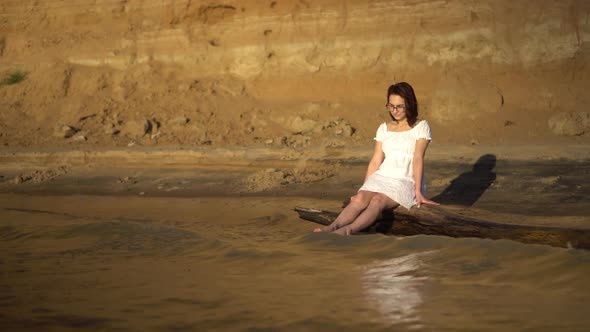 An Attractive Young Woman Is Sitting on a Log By the River. A Girl in a White Dress Splashes Her