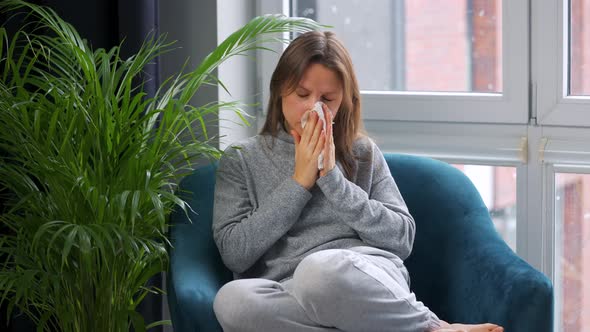 Unhealthy Woman Sits in a Chair and Sneezes or Blows Her Nose Into a Napkin Because She Has a Cold