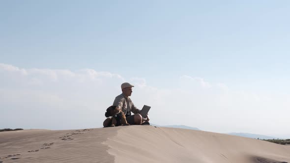 Male traveler sitting on sandy dune with dog and using laptop during vacation in sunny day