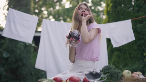 Slim Young Rural Woman Tasting Delicious Berry Sitting on Backyard with Clean Laundry and Harvest on