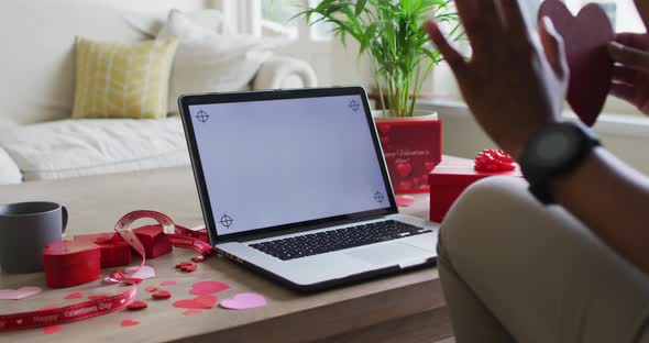 Hands of biracial woman holding heart and having valentine's video call on laptop