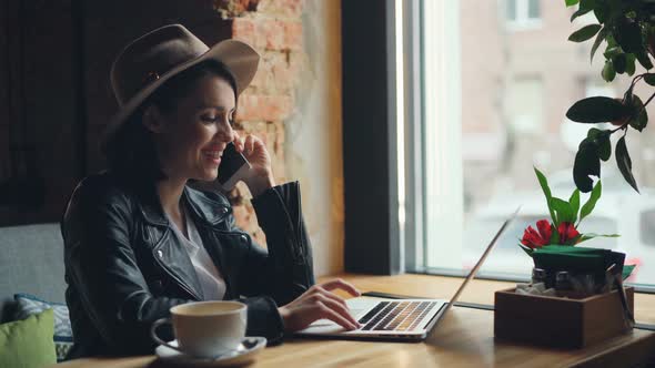 Happy Young Woman Talking on Mobile Phone and Using Laptop Typing in Cafe