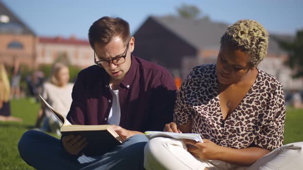 Diverse Classmates Preparing for Exam with Books and Workbook Sitting on Campus Lawn