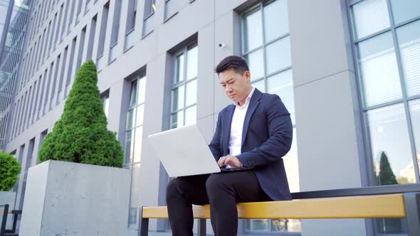 Male asian businessman working with laptop online outdoors sitting on bench in downtown 