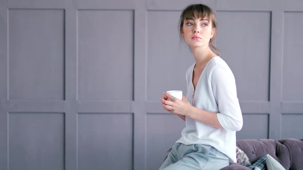 Casual Creative Young Woman Drinking Beverage Holding Cup Sitting on Armrest of Sofa