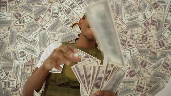 Black man with glasses is lying on a pile of money and waving dollar bills in hands.