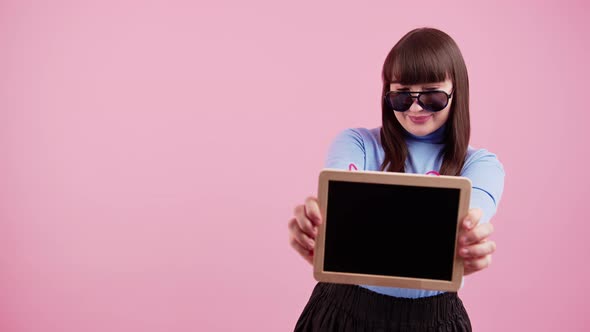 Cute Hipster European Brunette Teenager in Sunglasses Showing Mockup Blackboard to Camera Over Pink