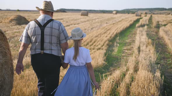 Grandfather and Granddaughter Walking Across the Field with Haystacks. Farmer Grandfather Teaches