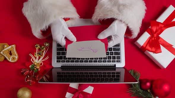Top View Santa Hands in White Gloves Are Typing on Keyboard Laptop By Red New Year Decorated Table