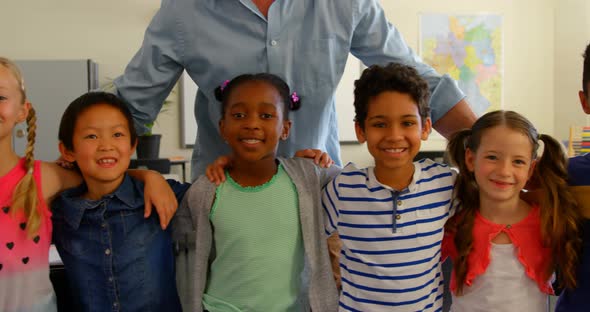 Front view of happy multi-ethnic schoolkids with Caucasian male schoolteacher standing in classroom