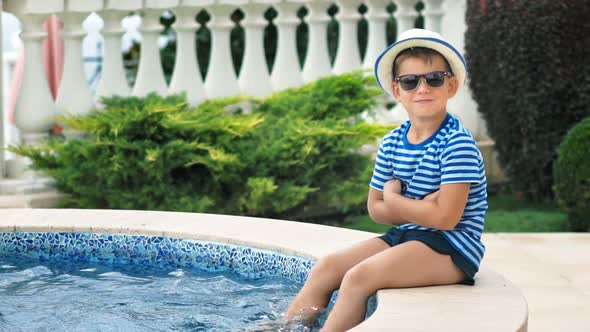 Slow Motion Little Boy in Sunglasses and Hat Sitting on Edge of Swimming Pool Playing Legs in Water