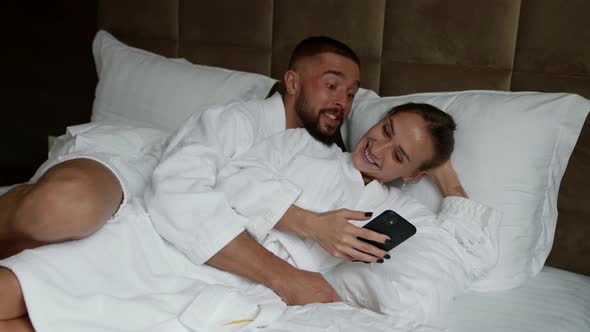 a Couple in White Coats in Bed Looking at the Phone and Smiling