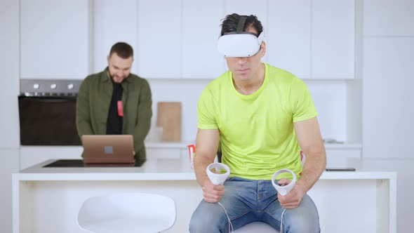 Absorbed Man Using VR Headset and Touch Controllers with Blurred Guy Surfing Internet on Laptop at