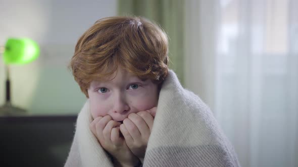 Portrait of Terrified Caucasian Redhead Boy Sitting Indoors Covered with Blanket and Looking Around