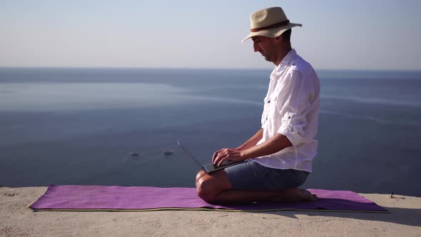 A Digital Nomad a Man in a Hat a Businessman with a Laptop Does Yoga on the Rocks By the Sea at