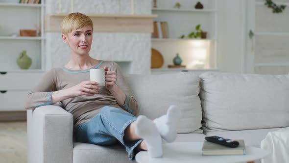 Dreamy Carefree Happy Woman Sitting on Sofa Drinking Morning Coffee or Warm Tea at Home Smiling