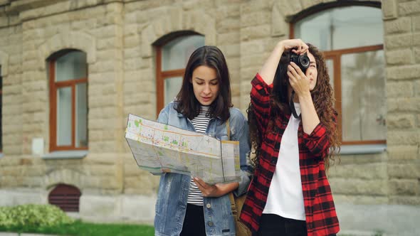 Good-looking Woman with Dark Hair Is Looking at Map While Her Female Friend Is Taking Pictures with