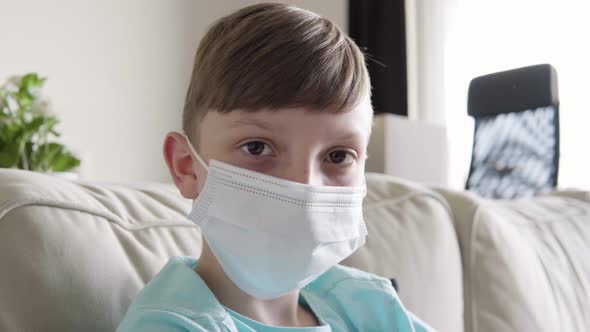 A Young Boy in a Face Mask Looks at the Camera As He Sits on a Couch at Home - Face Closeup