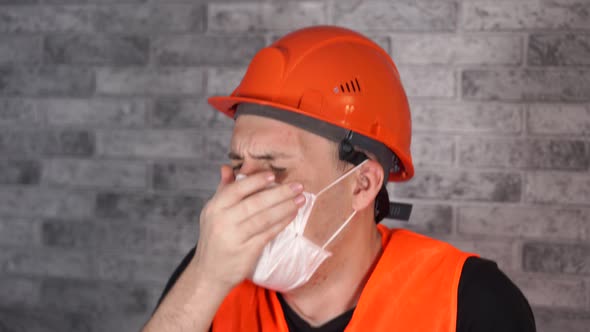 Male Construction Worker in Overalls and Medical Mask Coughing on Face Background of Gray Brick Wall