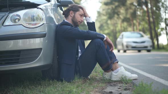 Side View Hopeless Young Man Sitting at Car Breakdown Lights on with Vehicle Passing on Suburban