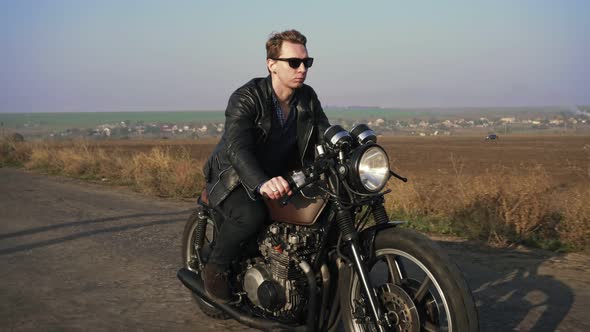 Side View of a Stylish Cool Young Man in Sunglasses and Leather Jacket Driving His Chopper on a