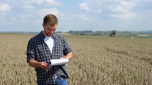 Happy Young Farmer with Notebook Standing on Wheat Field While Combine Harvester Working in