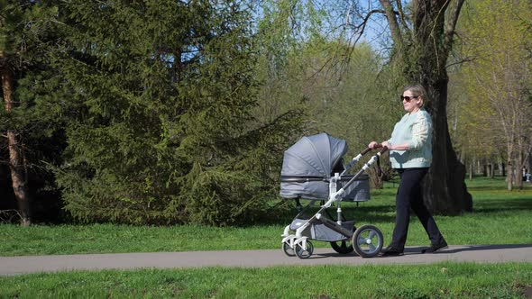 Grandma Walks with Stroller on Road in City Park in Spring