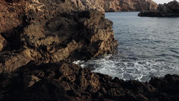 Rough sea waves crashing on a rocky shore. Dark brown cliffs by the sea in the sunset.