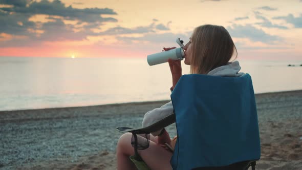 Young Woman Drinking From Thermos and Sitting on Camping Chairs on the Beach