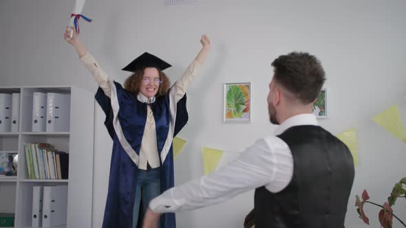 Young Cheerful Girl in Academic Clothes Jumps on Couch Rejoices in Diploma and Jumps Into Arms of