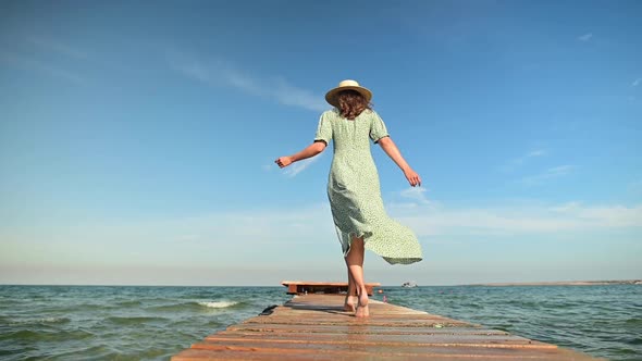 A Young Woman in a Green Summer Dress and a Straw Hat in Slow Motion Walks Along a Wooden Pier in