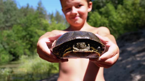 Boy Holds Turtle in Arms and Smiles on Background of River with Green Vegetation