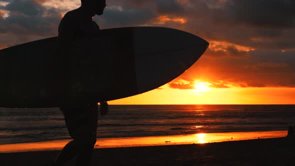 Young Surfer Man Walking on the Beach Holding Surf Board with Amazing Sunset on Background