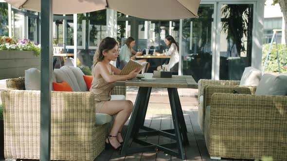 Attractive Girl Reading Interesting Book in Outdoor Cafe Relaxing Sitting at Table Alone