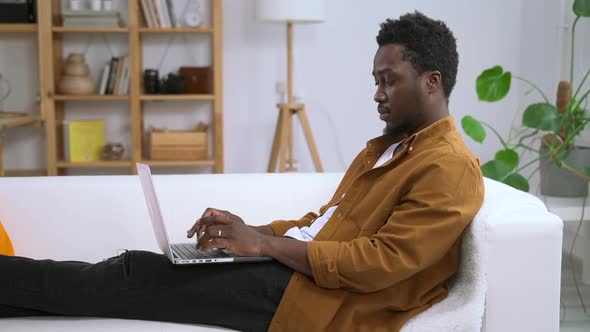 African Man with Computer Sits on Sofa Against Background of Home Interior Spbas