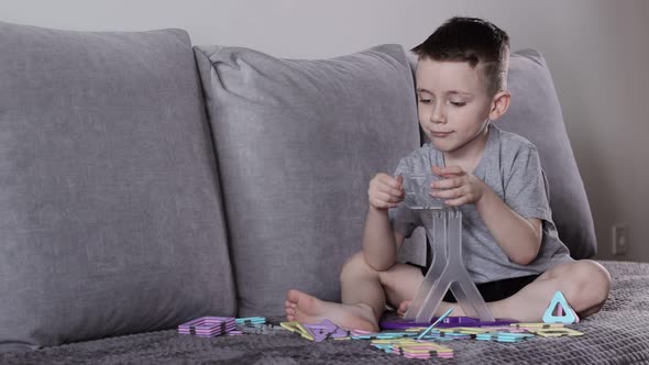 A boy in a gray T-shirt sits on a bed near a white wall and making Ferris wheel with constructor