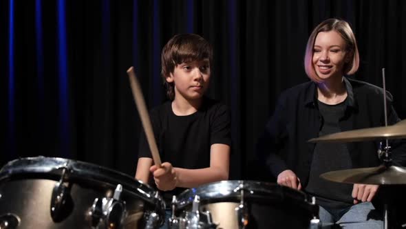 Young Caucasian Woman Teaches a Boy to Play the Drums in the Studio on a Black Background