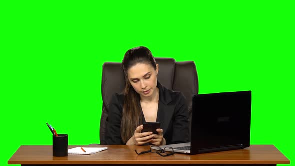Business Woman Sitting at a Desk Writes a Message on a Cell Phone with Rage. Green Screen. Studio
