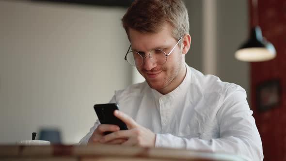 Young Man with Glasses is Resting in Cafe and Chatting Online in Social Networks By Mobile Phone