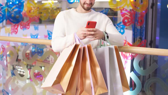 Portrait of Happy Shopaholic with Paper Bags in Hands Using Smartphone