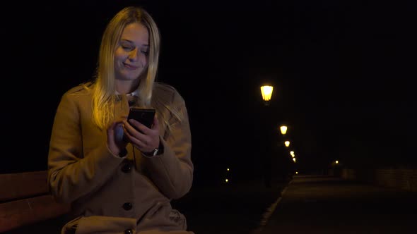 A Young Woman Sits on a Bench and Works on a Smartphone with a Smile in an Urban Area at Night