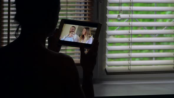 a Woman Standing Against the Light at a Window with Blinds, She Is Talking on a Video Link on a