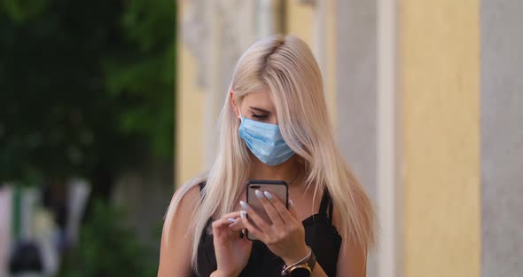 Young Blonde Woman in Protective Medical Mask Walks Down To the Street Uses Phone Texts Scrolls