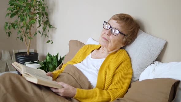 Sleeping Senior Woman Holding Book And Taking Nap In Bed.