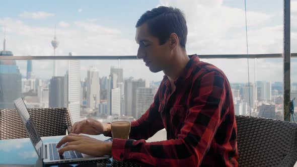 Confident Business Man Working on a Laptop and Drinking Coffee in Cafe on the Roof of a Skyscraper