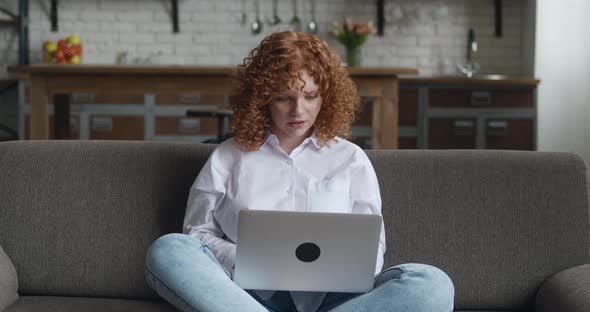 Happy Young Redhead Woman Sitting in Cozy Living Room on Couch Holding Using Laptop