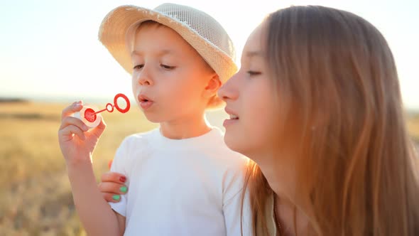 Teenage Sister Stands and Smiles Next to Little Brother Who Blows and Gets Soap Bubbles in Outdoor