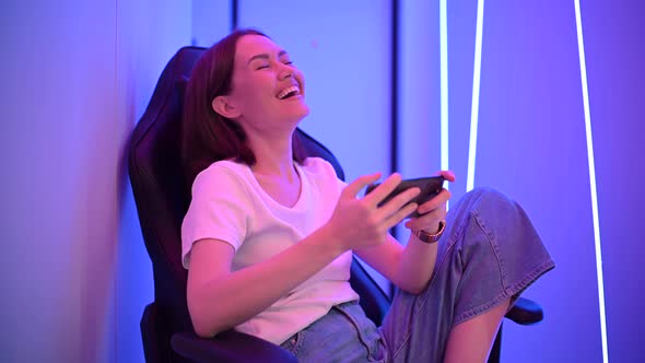Girl Relaxing Playing Games on Mobile Phone and Sitting on a Gaming Chair. Room with Colorful Neon