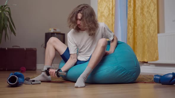 Wide Shot of Weak Funny Retro Man Sitting on Bag Chair Training Biceps with Dumbbell