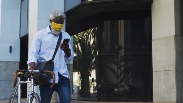 African american senior man wearing face mask with bicycle using smartphone in corporate park